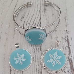May include: A set of three silver snowflake jewelry pieces. A silver bangle bracelet with a blue snowflake charm, a silver pendant with a blue snowflake charm, and a silver pendant with a blue snowflake charm surrounded by rhinestones.