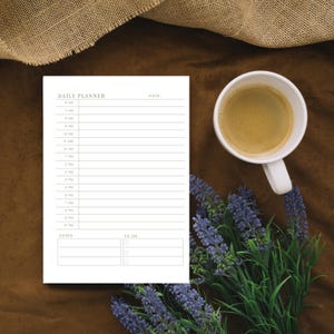 Puede incluir: Un planificador diario blanco con el texto "DAILY PLANNER" y una taza de café sobre una superficie marrón. El planificador tiene un horario de 6 AM a 10 PM y una sección "TO-DO". Un ramo de flores moradas y una tela de arpillera también están en la imagen.