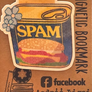 May include: A magnetic bookmark featuring a Spam can design with a sandwich illustration. The bookmark has the text "SPAM" in blue letters and is decorated with blue flowers. The words "MAGNETIC BOOKMARK" are printed vertically.