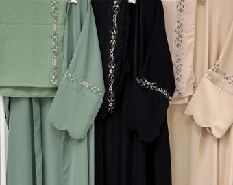 May include: Several modest outfits hang on white hangers. The outfits are in various colors, including sage green, black, and beige. Each outfit features floral embroidery with pink accents. The sleeves and edges of the garments are adorned with delicate floral patterns.