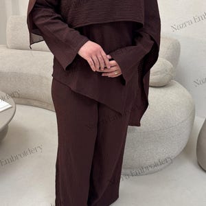 May include: A dark brown ensemble featuring a long-sleeved top with a cape-like overlay and matching wide-leg pants. The outfit includes a head covering and is accessorized with a gold brooch. The fabric has a textured appearance.