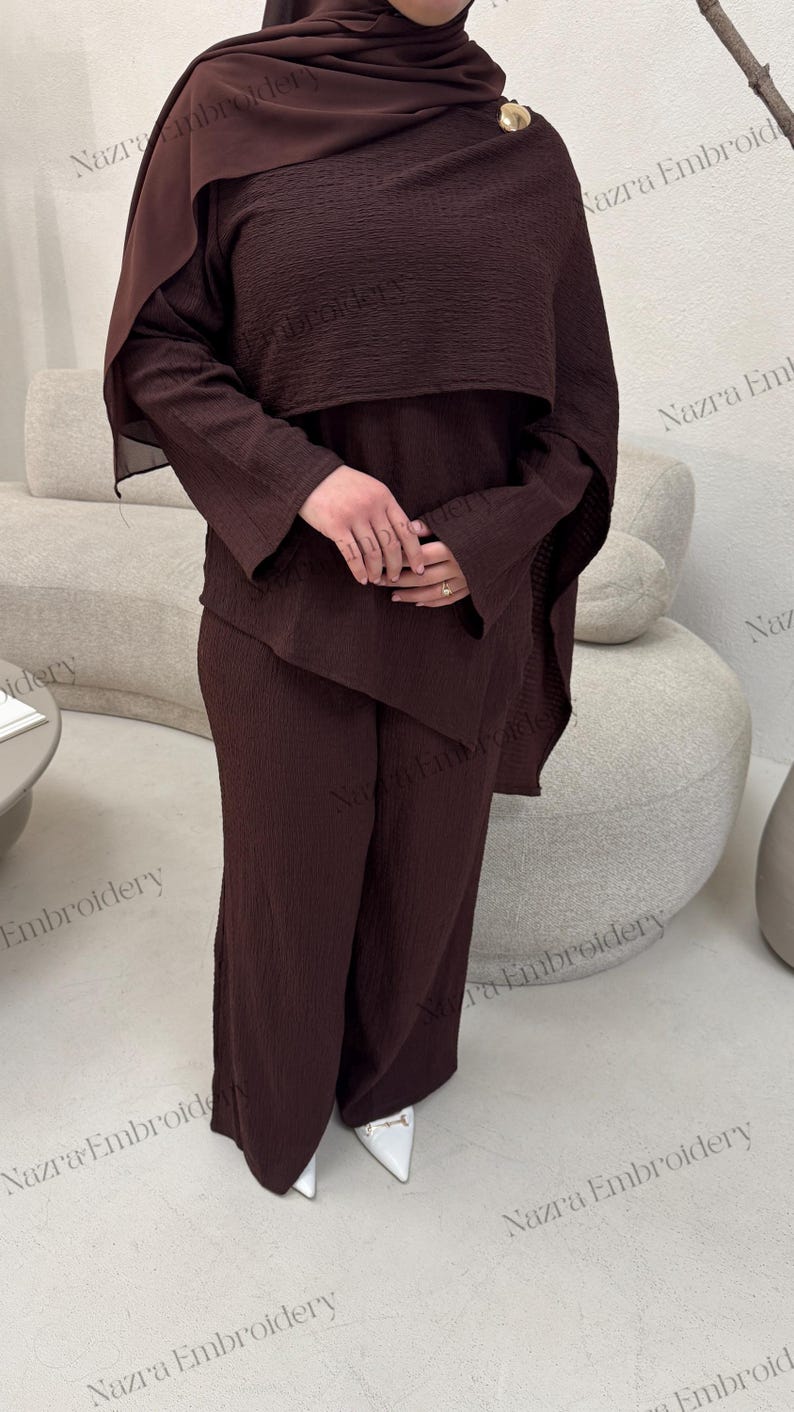 May include: A dark brown ensemble featuring a long-sleeved top with a cape-like overlay and matching wide-leg pants. The outfit includes a head covering and is accessorized with a gold brooch. The fabric has a textured appearance.