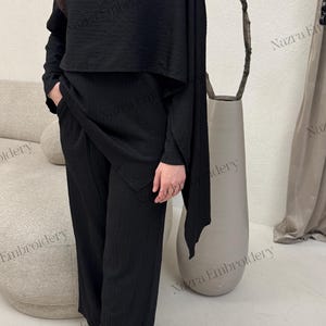 May include: Black two-piece outfit featuring a textured top with a gold shoulder detail and wide-leg pants. The outfit includes a matching top and pants set. The model is wearing white pointed-toe heels.