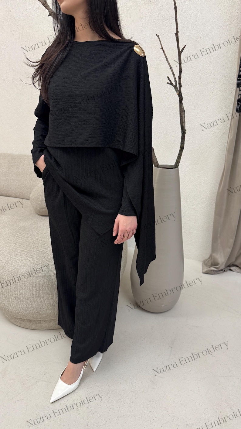 May include: Black two-piece outfit featuring a textured top with a gold shoulder detail and wide-leg pants. The outfit includes a matching top and pants set. The model is wearing white pointed-toe heels.