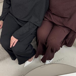 May include: Two matching sets of women's clothing. One set is black, the other is a deep burgundy. Each set includes a long-sleeved top with a gold button detail and matching pants. White shoes are visible.