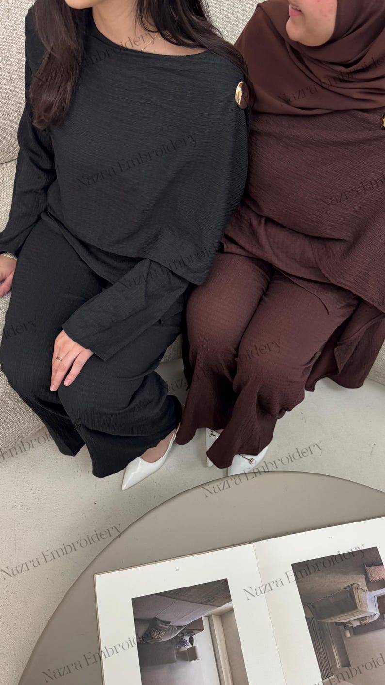 May include: Two matching sets of women's clothing. One set is black, the other is a deep burgundy. Each set includes a long-sleeved top with a gold button detail and matching pants. White shoes are visible.