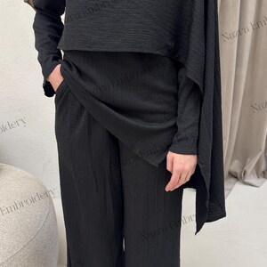 May include: A black two-piece outfit featuring a textured fabric. The ensemble includes wide-leg pants and a top with long sleeves and a draped cape detail, fastened with a gold button. White pointed-toe heels complete the look.