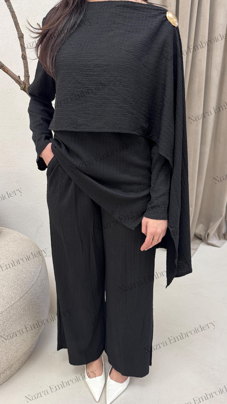May include: A black two-piece outfit featuring a textured fabric. The ensemble includes wide-leg pants and a top with long sleeves and a draped cape detail, fastened with a gold button. White pointed-toe heels complete the look.
