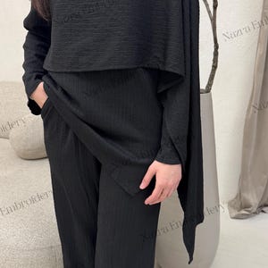 May include: A black two-piece outfit featuring a long-sleeved top with a cape-like overlay and wide-leg pants. The top has a round neckline and a gold-colored shoulder detail. The fabric has a textured, ribbed appearance. The outfit is suitable for formal or casual wear.