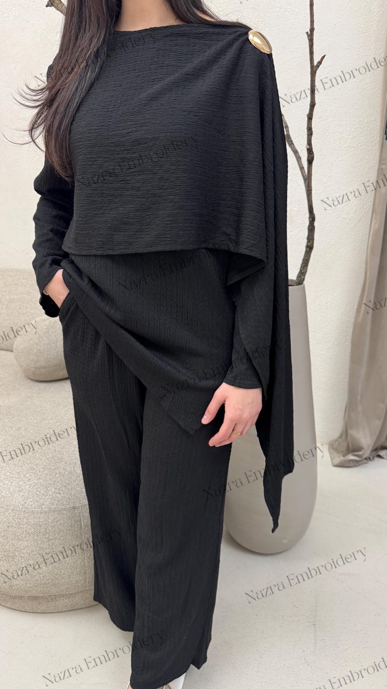 May include: A black two-piece outfit featuring a long-sleeved top with a cape-like overlay and wide-leg pants. The top has a round neckline and a gold-colored shoulder detail. The fabric has a textured, ribbed appearance. The outfit is suitable for formal or casual wear.
