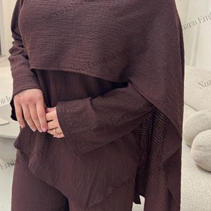 May include: A brown two-piece outfit with a textured fabric. The ensemble includes a long-sleeved top with a draped overlay and matching pants. A gold-colored button accents the top. The outfit is suitable for modest wear.