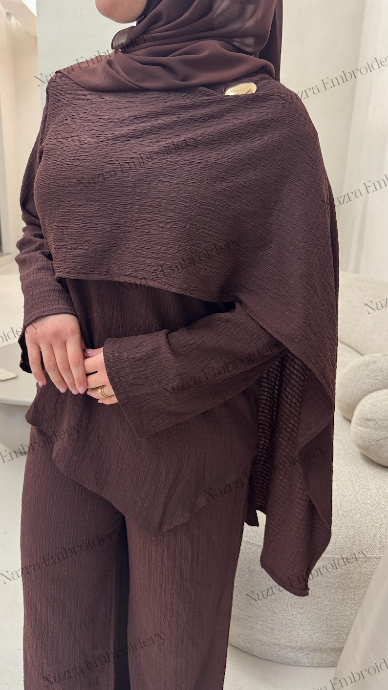 May include: A brown two-piece outfit with a textured fabric. The ensemble includes a long-sleeved top with a draped overlay and matching pants. A gold-colored button accents the top. The outfit is suitable for modest wear.