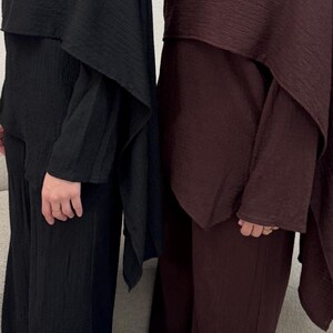 May include: Two matching outfits in the image. One is black, the other is a deep burgundy. Each outfit includes a long-sleeved top with a cape-like overlay and wide-leg pants. The fabric has a textured, crinkled appearance, suggesting a lightweight material.