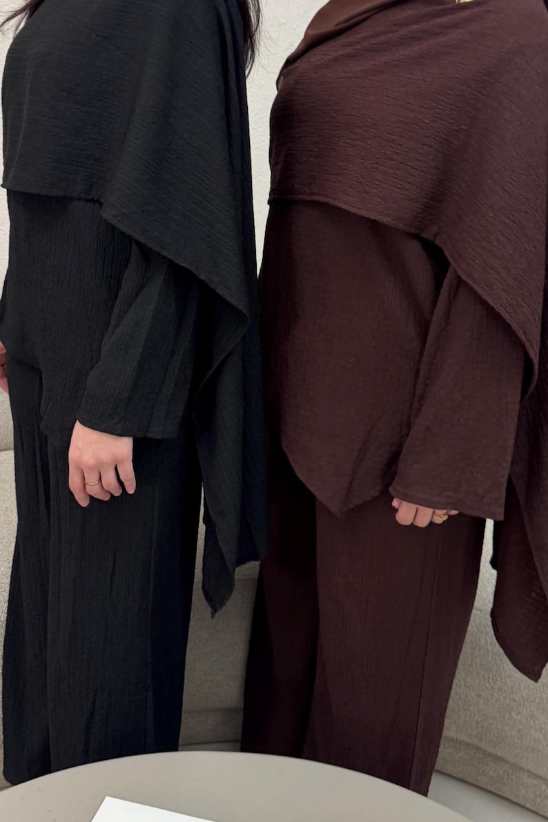 May include: Two matching outfits in the image. One is black, the other is a deep burgundy. Each outfit includes a long-sleeved top with a cape-like overlay and wide-leg pants. The fabric has a textured, crinkled appearance, suggesting a lightweight material.