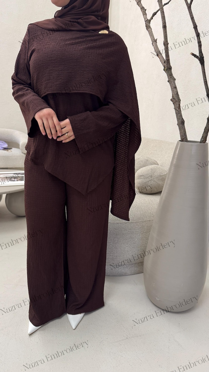 May include: A dark brown, textured two-piece outfit consisting of a long-sleeved top and wide-leg pants. The top has a cape-like overlay. The outfit is paired with a matching head covering. White pointed-toe shoes are visible.