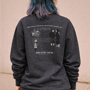 May include: Dark gray sweatshirt with a graphic on the back. The graphic includes text that reads "THE DEVIL AND GOD ARE RAGING INSIDE ME." and "BRAND NEW." There are also illustrations of figures and flowers.