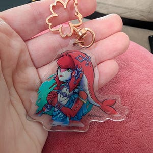 May include: A clear acrylic keychain featuring a character with red hair and a blue outfit. The keychain has a gold-colored flower-shaped clasp and a round ring. The character design is a stylized illustration with a detailed face and clothing.