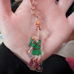 May include: A clear acrylic keychain featuring a cartoon illustration of a character in a green tunic and hat, holding a torch. The keychain has a gold-colored chain and ring. The character is reaching upwards, with a small fairy nearby.