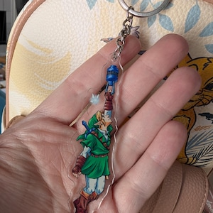 May include: A clear acrylic keychain featuring a cartoon illustration of a character in a green tunic, brown boots, and a blue hat. The character is holding a blue object. The keychain is attached to a silver metal ring.