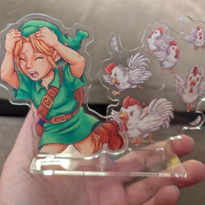 May include: A clear acrylic stand-up featuring a cartoon illustration of a character in a green tunic, with blonde hair, looking distressed, surrounded by cartoon chickens. The chickens have red combs and are in various poses.
