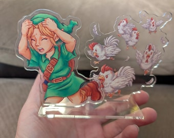 May include: A clear acrylic stand-up featuring a cartoon illustration of a character in a green tunic, with blonde hair, looking distressed, surrounded by cartoon chickens. The chickens have red combs and are in various poses.