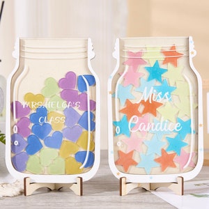 May include: Two white wooden mason jar shaped containers with a stand. One jar is filled with colourful hearts and says "Mrs. Helga's Class". The other jar is filled with colourful stars and says "Miss Candice".
