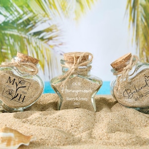 May include: Three glass jars with cork lids and twine tied around the necks. The jars are filled with sand and have different designs. One jar has a heart shape with the text "M&H July 2023", another jar has a star shape with the text "Our Honeymoon Barry & Jenny 2023", and the third jar has a heart shape with the text "Behind 05.04.2023".