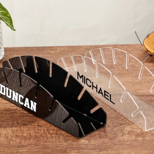 Puede incluir: Dos soportes para bate de béisbol de acrílico, uno negro y otro transparente, con los nombres "Duncan" y "Michael" impresos en blanco. Los soportes están sobre una superficie de madera.
