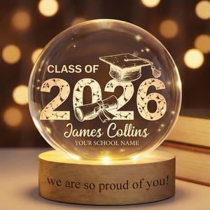 May include: A clear glass graduation sphere with the text "CLASS OF 2026" and a mortarboard and diploma design. The sphere sits on a wooden base with the words "we are so proud of you!" and is illuminated from within.