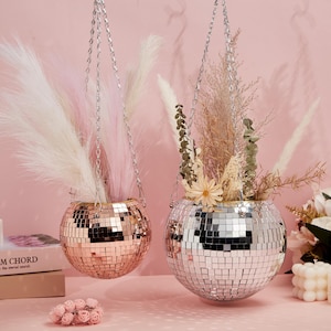Pode incluir: Dois vasos suspensos em forma de bola de discoteca com flores secas. O vaso à esquerda é dourado-rosado e o vaso à direita é prateado.