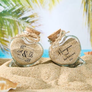 Pode incluir: Dois frascos de vidro em forma de coração cheios de areia, um com a etiqueta "Honeymoon Sand Barry & Jenny 2023" e o outro "A&S May 2023".