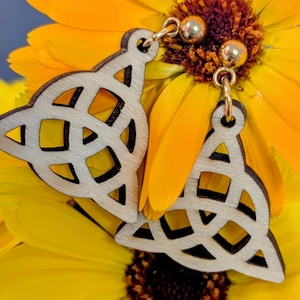 May include: Light wooden Celtic knot earrings with gold-coloured ear wires. The earrings have a geometric design and are displayed on a bright yellow flower with a dark brown centre. The background is a soft blue.