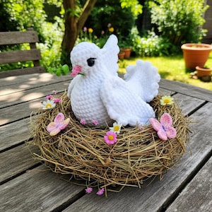 Könnte beinhalten: Eine handgefertigte weiße Häkeltaube mit rosa Schnabel und schwarzen Augen, die einen grünen Zweig hält, sitzt in einem gewebten Nest. Das Nest ist mit kleinen rosa und gelben Blumen und zwei rosa Häkel-Schmetterlingen verziert.