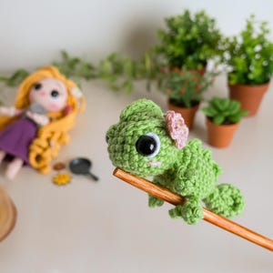 May include: A small, green, crocheted chameleon figure with large black eyes and a light pink flower detail on its head. The figure is perched on a wooden stick. In the background, a doll with long yellow hair and small potted plants are visible.