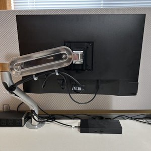 May include: A black computer monitor is mounted on a silver adjustable arm. The monitor is connected to a black box with a variety of cables. The monitor is mounted on a white desk.