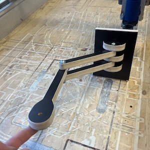 Peut inclure: Un bras articulé noir et bois avec trois articulations. Le bras est fixé à une base noire et est fait de contreplaqué.