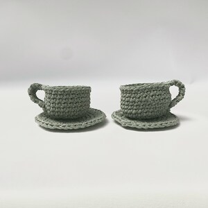 May include: Two crocheted teacups and saucers in a light green color. The cups and saucers are made of yarn and have a textured surface.