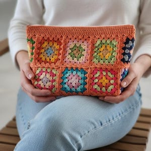 Puede incluir: Un neceser de ganchillo color melocotón con cierre de cremallera. El neceser presenta un diseño de patchwork de cuadrados de la abuela coloridos en tonos rosa, azul, verde y amarillo. La bolsa hecha a mano se sostiene en el regazo de una persona.