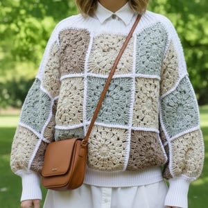 May include: A long-sleeve crochet jumper featuring a patchwork of square motifs in shades of sage green, beige, and cream, worn over a white collared shirt. A tan leather crossbody bag with a flap closure hangs across the front of the jumper.