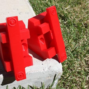 May include: Two red plastic blocks with a series of holes and slots. The blocks are designed to be used together to create a specific shape.