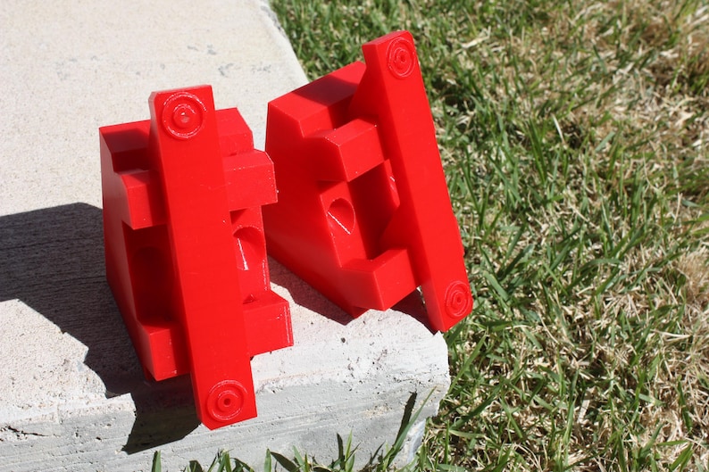 May include: Two red plastic blocks with a series of holes and slots. The blocks are designed to be used together to create a specific shape.
