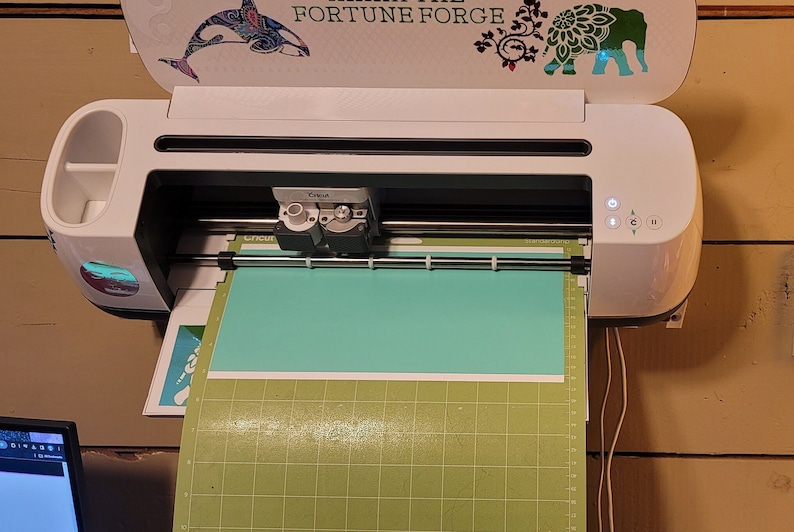 May include: A white Cricut Explore Air 2 cutting machine with a teal mat and a green cutting mat. The machine is on and the lid is open. The machine is labelled "Cricut" and has a small screen with a few buttons.