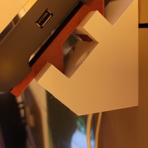 May include: A white wall mount with a red and white base, designed to hold a speaker. The speaker is light grey and white and has a USB port.