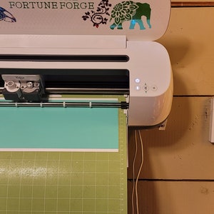 May include: A white Cricut Explore Air 2 cutting machine with a light blue cutting mat and a green cutting mat. The machine is mounted on a wall and has a sticker on the top that says "Fortune Forge".