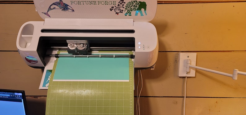May include: A white Cricut Explore Air 2 cutting machine with a light blue cutting mat and a green cutting mat. The machine is mounted on a wall and has a sticker on the top that says "Fortune Forge".