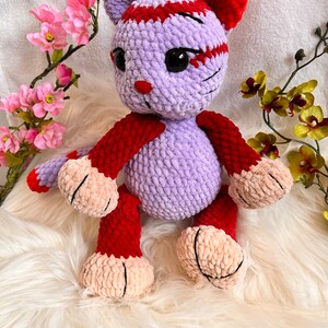 May include: A crocheted plush toy in the shape of a cat. The cat is purple with red accents and has black eyes. The cat is sitting on a white fluffy surface.