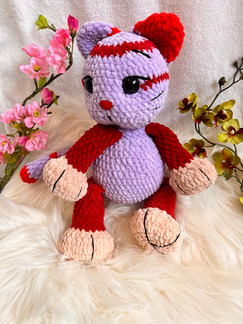 May include: A crocheted plush toy in the shape of a cat. The cat is purple with red accents and has black eyes. The cat is sitting on a white fluffy surface.