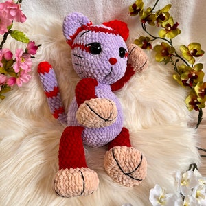 May include: A crocheted plush toy cat with a purple body, red and white stripes, and pink paws. The cat is sitting on a white fluffy surface.