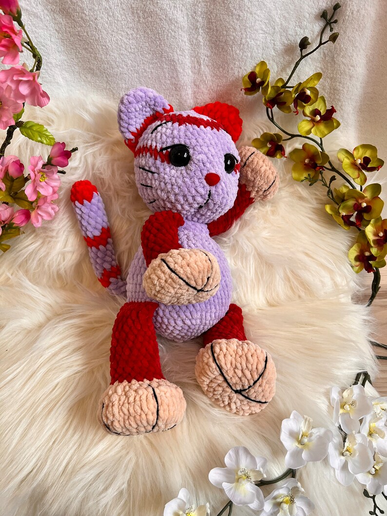 May include: A crocheted plush toy cat with a purple body, red and white stripes, and pink paws. The cat is sitting on a white fluffy surface.