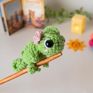 May include: A small, green, crocheted chameleon plush toy with a textured surface, large black plastic eyes, and a light pink flower detail on its head. The creature is perched on a smooth, light brown wooden stick, showcasing its detailed yarn stitches.